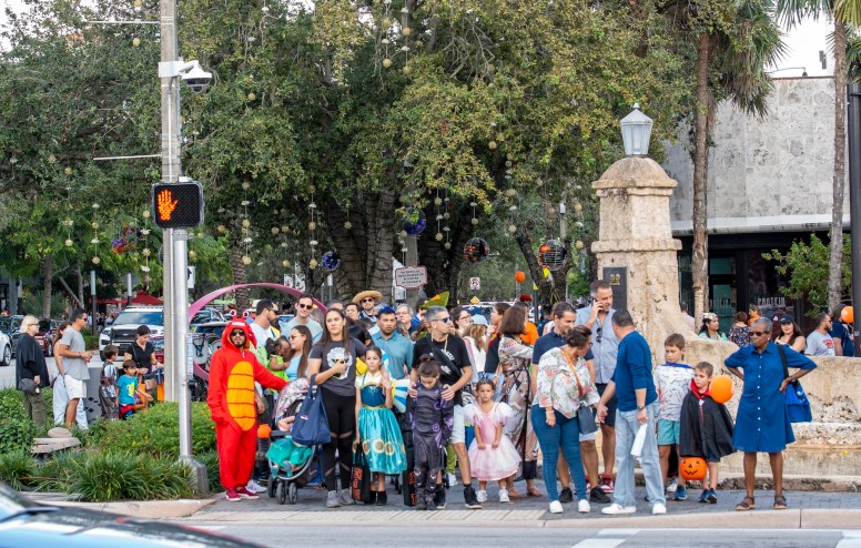 a big group of trick-or-treaters in costume and their parents wait to cross the street at a crosswalk
