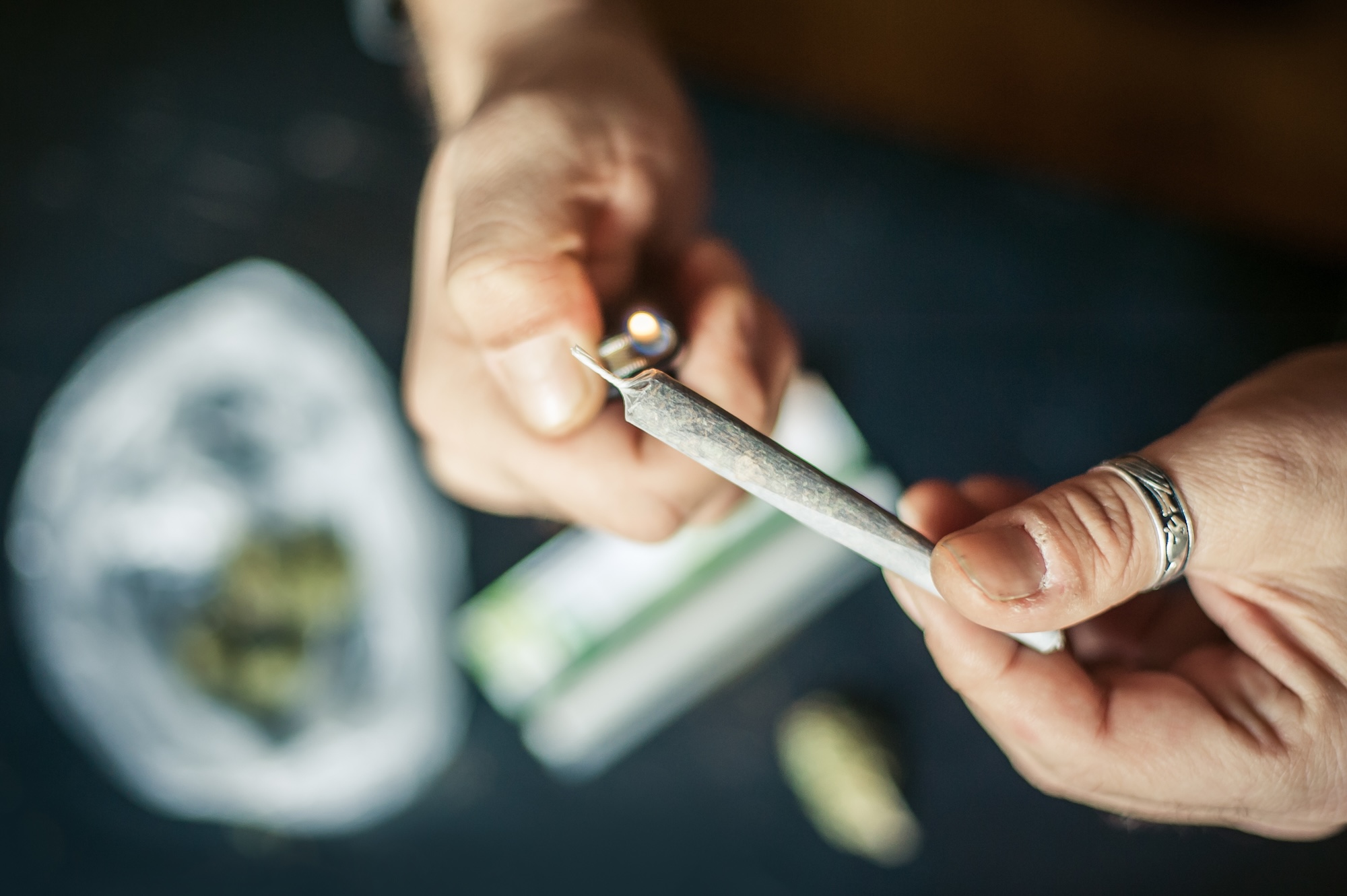 photo of a closeup of hands lighting a marijuana joint with marijuana on the table in a baggie on a table below