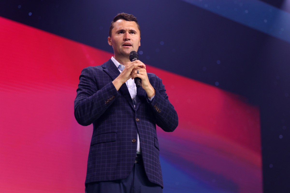Charlie Kirk in a suit holding a microphone in front of a navy and red backdrop