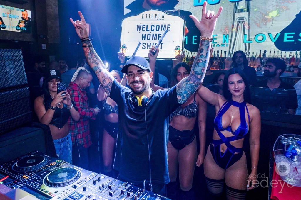 a DJ stands behind a booth flanked by bottle girls and fans