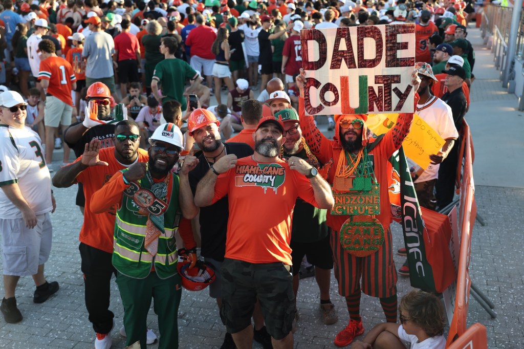 A group of sports fans dressed head-to-toe in UM gear and holding a sign reading, "Dade County" with the UM logo