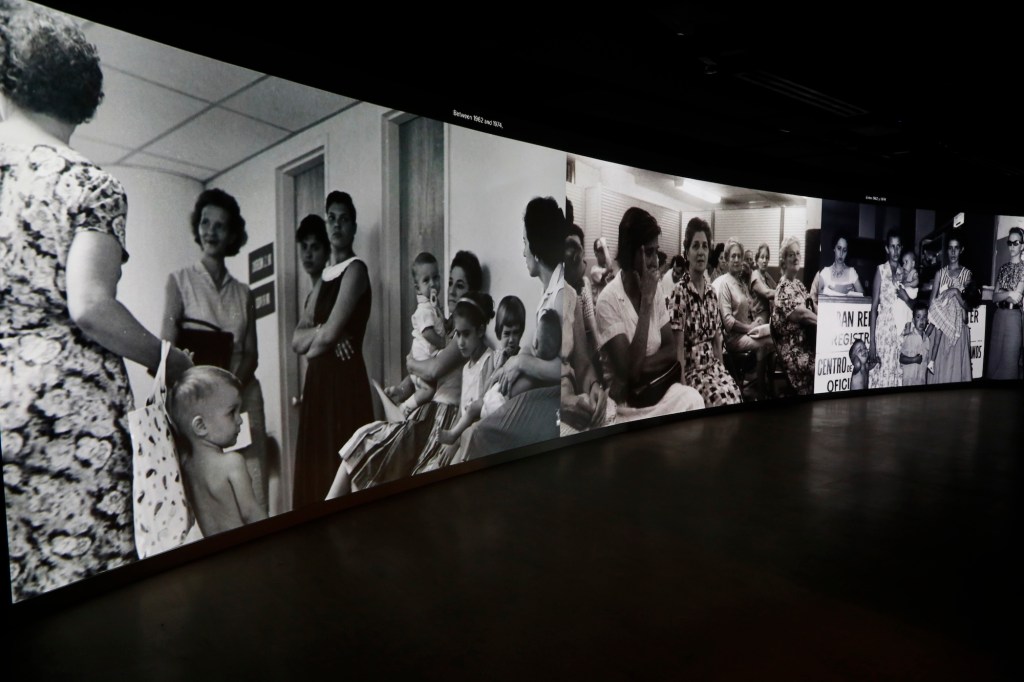 a curved museum screen on a wall shows black-and-white photos of Cuban refugees waiting to be processed in the 1960s