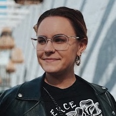 headshot of a bespectacled woman with short dark hair, wearing a black leather jacket over a black T-shirt emblazoned with white artwork