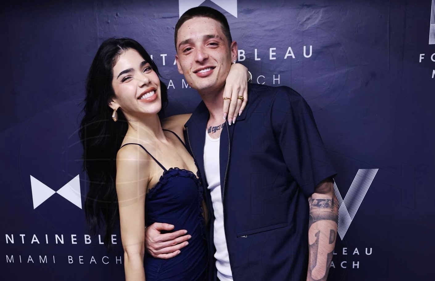Kenia Os and Peso Pluma hug and pose in front of a step and repeat backdrop reading "Fontainebleau Miami Beach