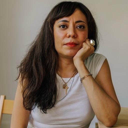 headshot of a young woman with dark hair wearing a sleeveless white top, seated in a wooden chair and resting her chin on her left hand with her elbow leaning on a wooden table