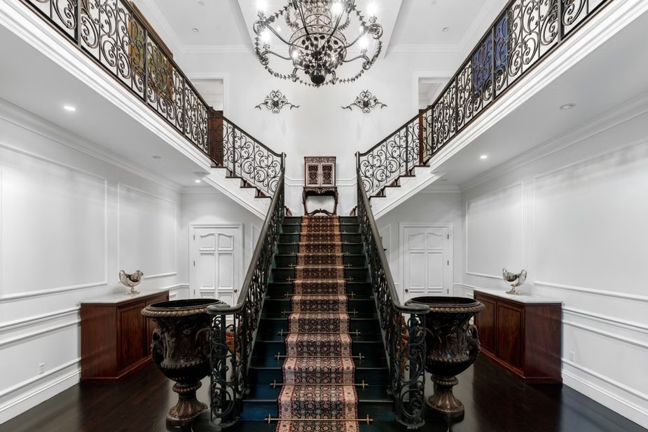 Depicted is a grand entryway with stark-white walls and an ornate staircase stretching underneath an elaborate chandelier.