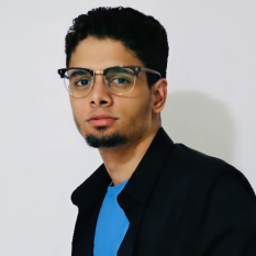 photo of a young, bespectacled man with a chin beard, wearing a black jacket with the collar turned up over a bright blue T-shirt