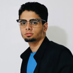 photo of a young, bespectacled man with a chin beard, wearing a black jacket with the collar turned up over a bright blue T-shirt