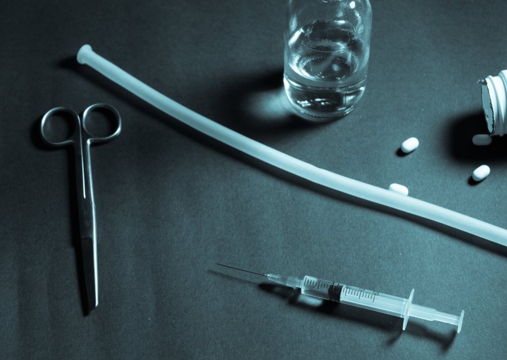 dim photo of a medical tray with instruments including scissors, a plastic tube, a syringe, a glass with clear liquid, and a bottle on its side with three pills spilled out