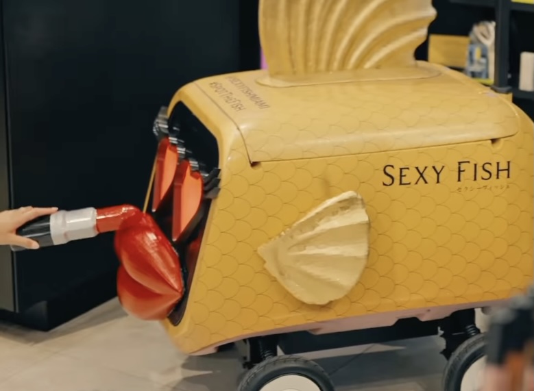 A food delivery robot with writing reading, "Sexy Fish" on the side. On the front, the robot has two heart-shaped eyes and red, plastic lips