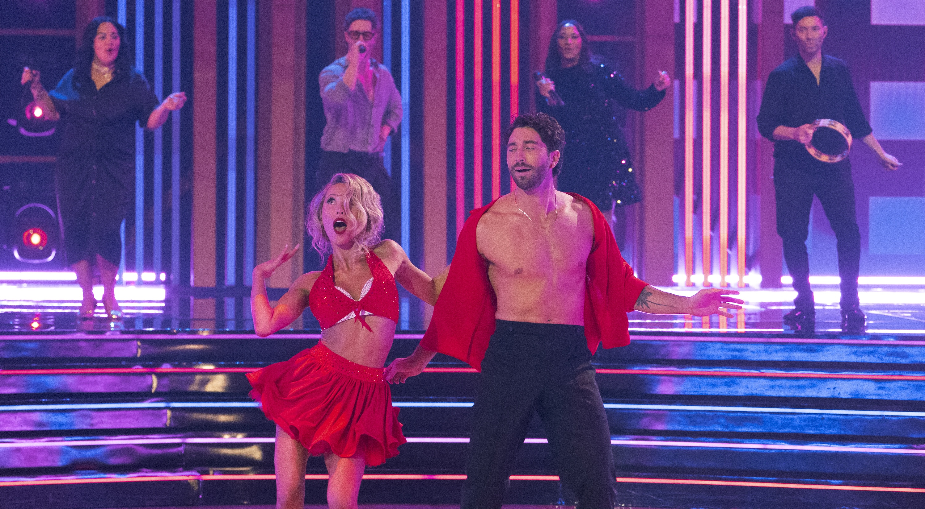 Photo of a woman and man dancing on a stage. The woman is on the left wearing a bedazzled red bralette and skirt with a bedazzled white bra underneath. The man, on the right, wears black pants and an open red, silk shirt. Behind them are two female singers, one male singer, and a man playing tambourine