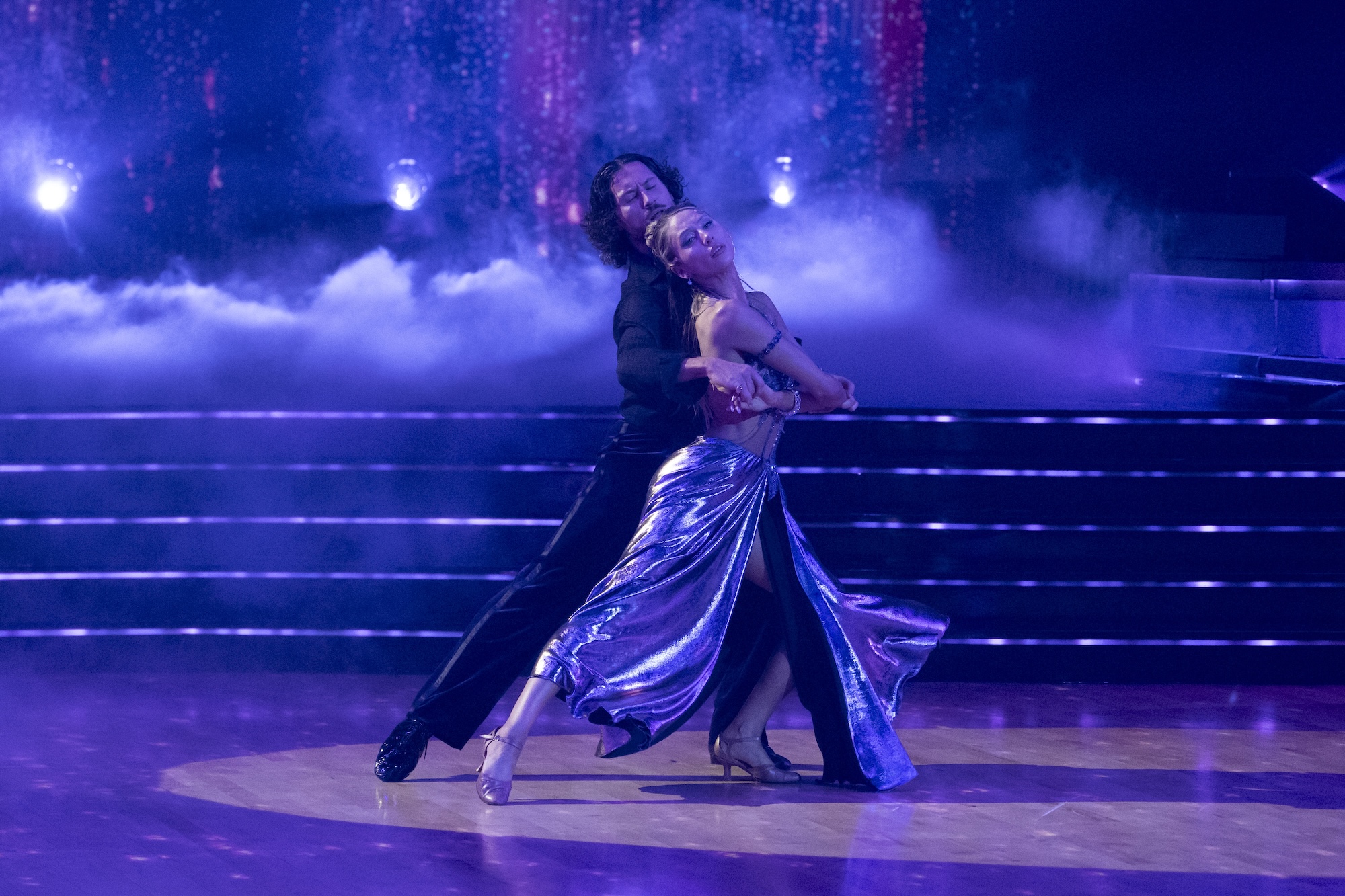 Photo of two dancers embracing while dancing on a ballroom floor. The man, wearing a black long-sleeved shirt and pants, embraces the woman, who wears a sparkling top and shimmering skirt