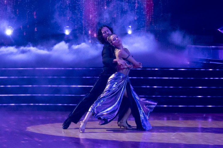 Photo of two dancers embracing while dancing on a ballroom floor. The man, wearing a black long-sleeved shirt and pants, embraces the woman, who wears a sparkling top and shimmering skirt
