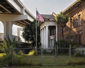 Photo of an abandoned school beneath a highway overpass behind a chainlink fence. The brick building has four columns at the entrance and an American flag flies in front of it.