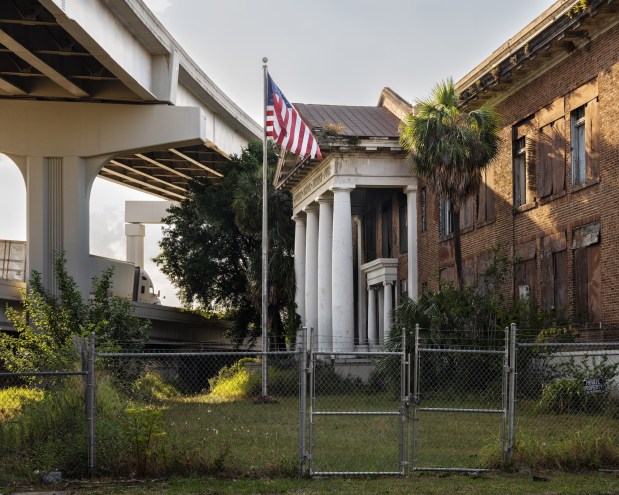Photo of an abandoned school beneath a highway overpass behind a chainlink fence. The brick building has four columns at the entrance and an American flag flies in front of it.
