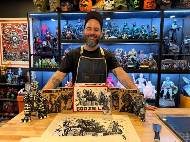 Photo of a bearded young artist wearing a baseball cap and apron inside his studio. Behind him are book shelves with dozens of action figures. In front of him is a print, a model, and wood stencils depicting Godzilla.