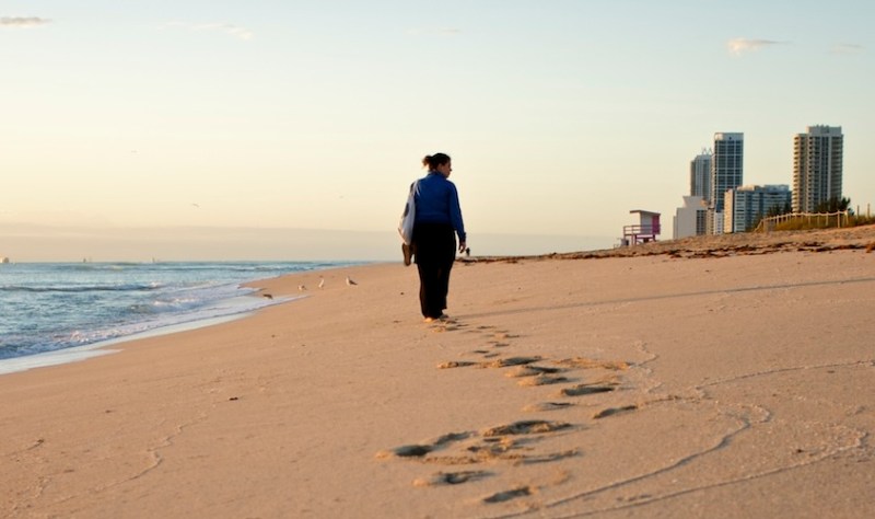 photo of a fully-clothed woman walking along the shore on the beach with high-rise buildings in the background on the right