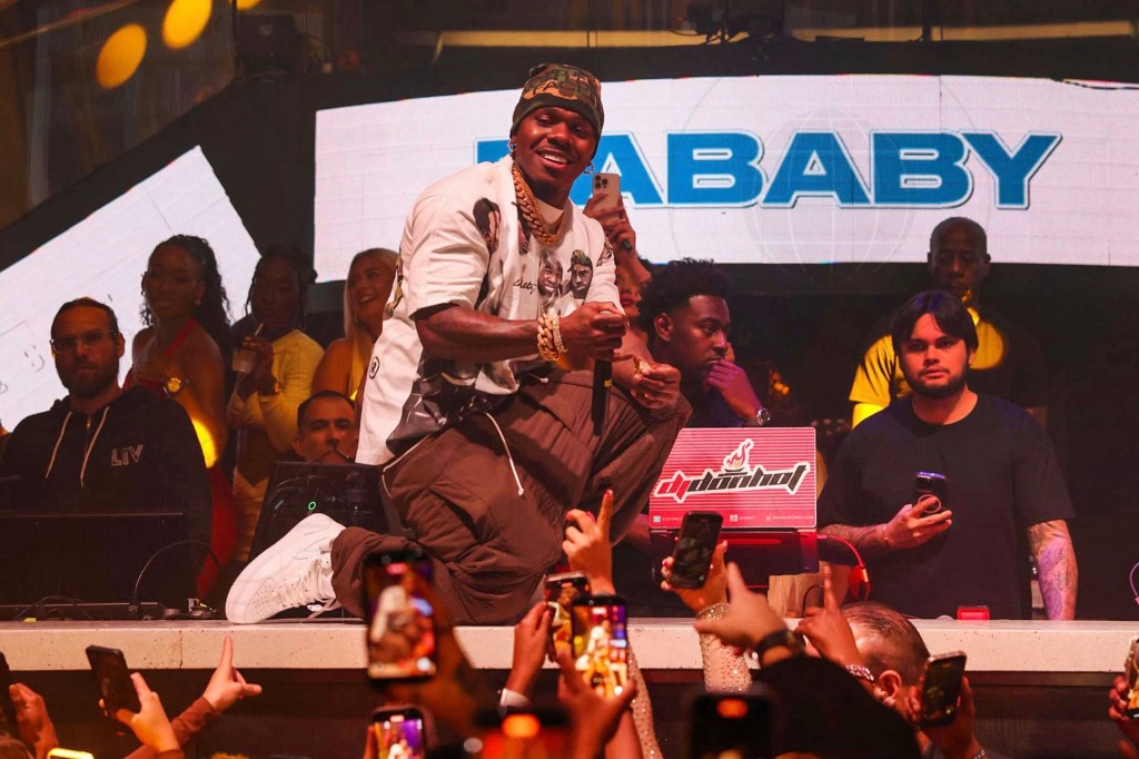 photo of rapper DaBaby kneeling on a DJ booth as he performs in front of an audience at a club