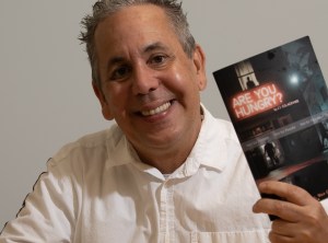 photo of author J.J. Colagrande in a white button-down T-shirt holding up a copy of his latest book, Are You Hungry. The cover is a dark image with two shadowy figures, with the name of the book in neon lights
