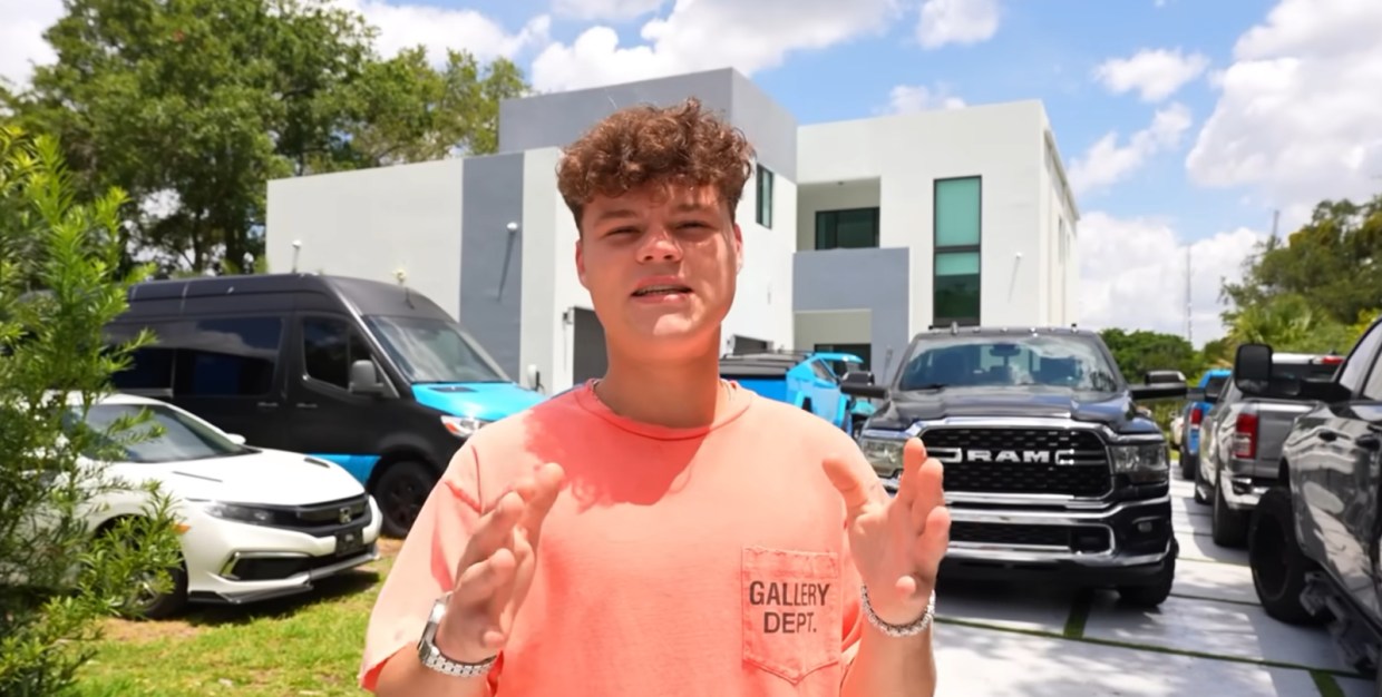 Jack Doherty who has a curly mop haircut, stands in front of his house speaking to the camera. He is wearing a salmon colored shirt.