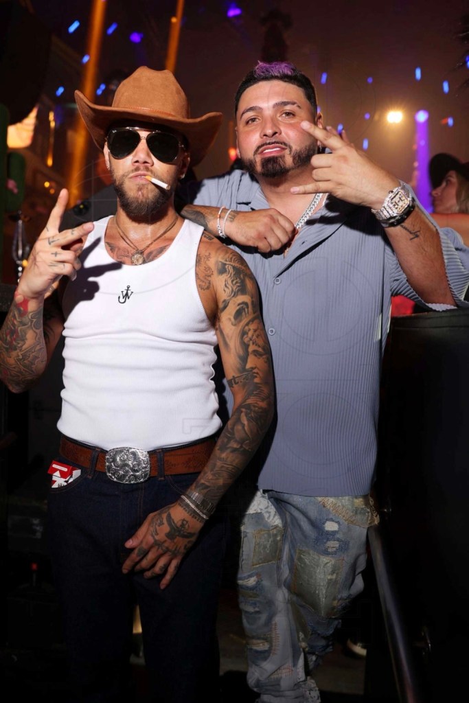Photo of a man wearing a cowboy hat with a cigarette hanging out of his mouth and posing next to another man in a baggy button-down and denim.