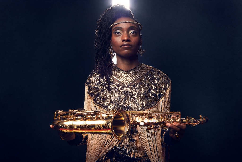 Photo of a woman wearing a golden dress and holding a saxophone.