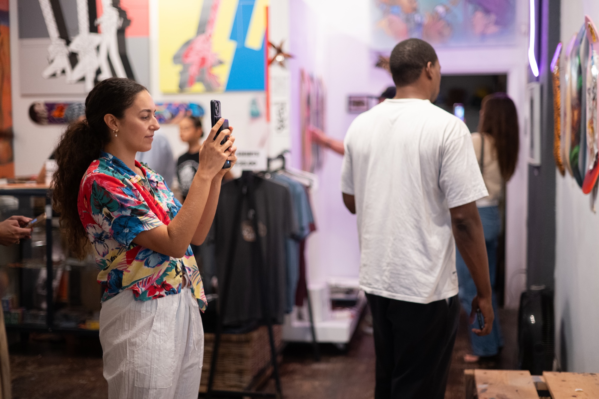 photo of a woman in a colorful shirt and white linen pants holding up her phone camera to take a photo of a wall in a gallery