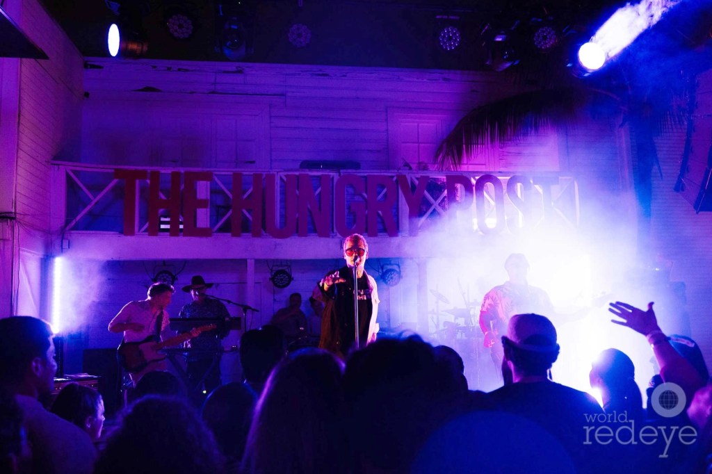 Photo of a band performing onstage in front of a crowd under purple lighting. A sign behind the band reads, "The Hungry Post"