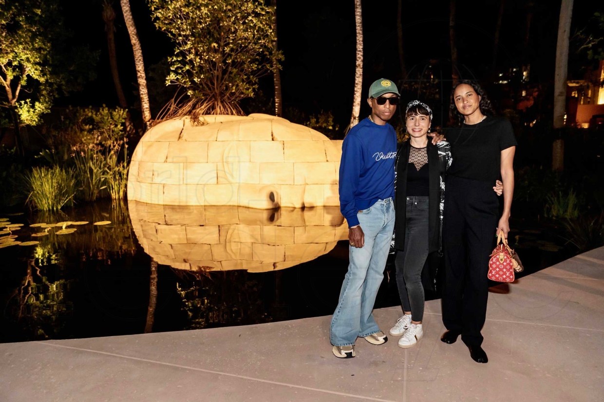photo of musician Pharrell Williams posing with two women in front of a glowing sculpture of a human head on its side