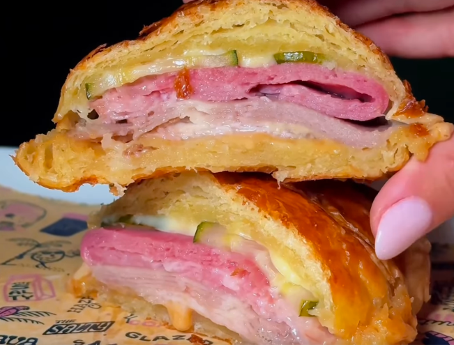 a croissant filled with ham, cheese, and pickles next to a woman's finger