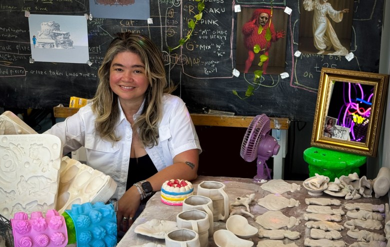 Photo of an artist in her studio. Behind her is a chalkboard with writing and posters. On the table beside her are clay scultures