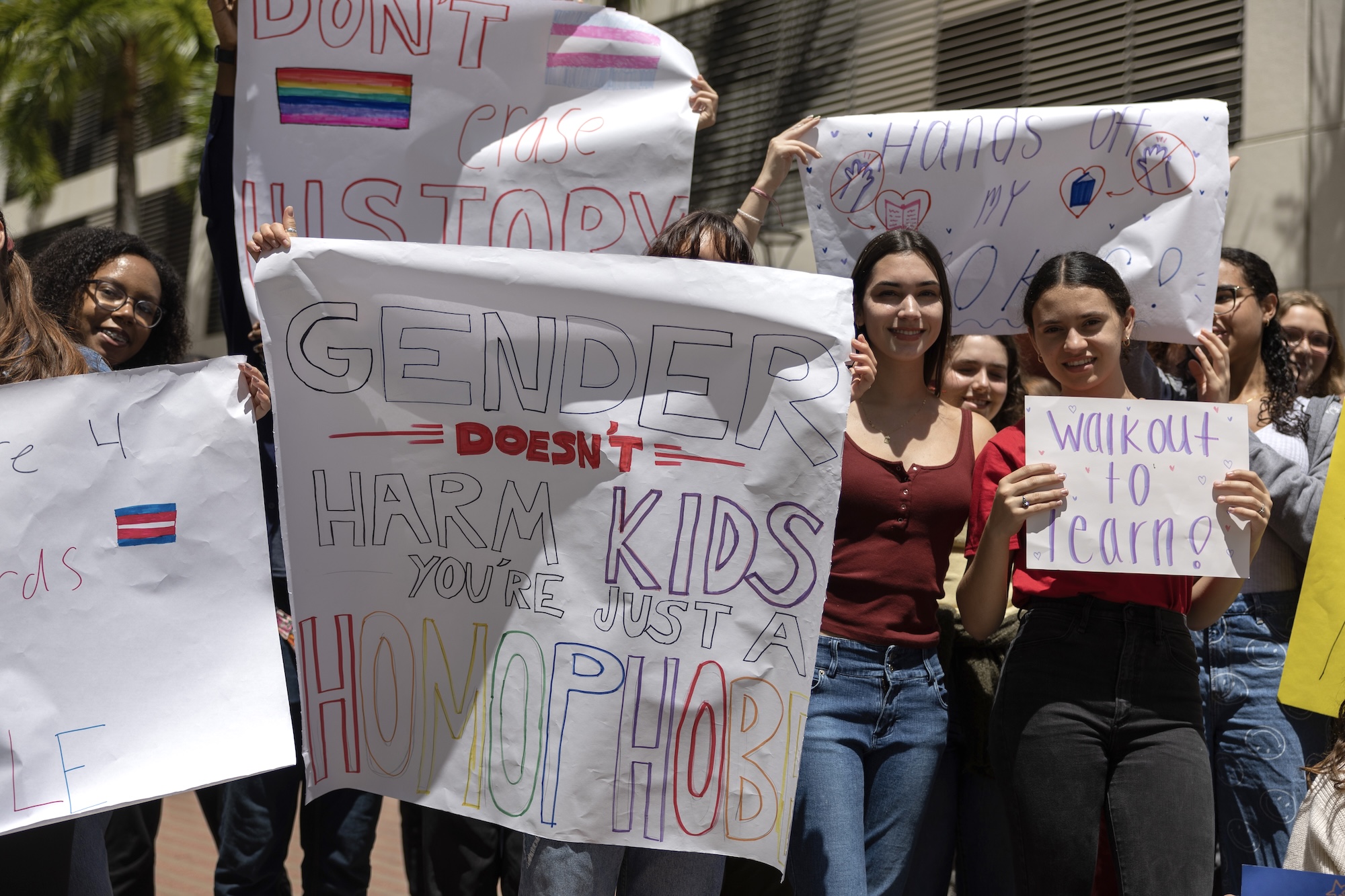 students hold up hand-drawn signs that read "Gender Doesn't Harm Kids You're Just a Homophobe," "Don't Erase History," "Walkout to Learn," and "Hands Off My Books"