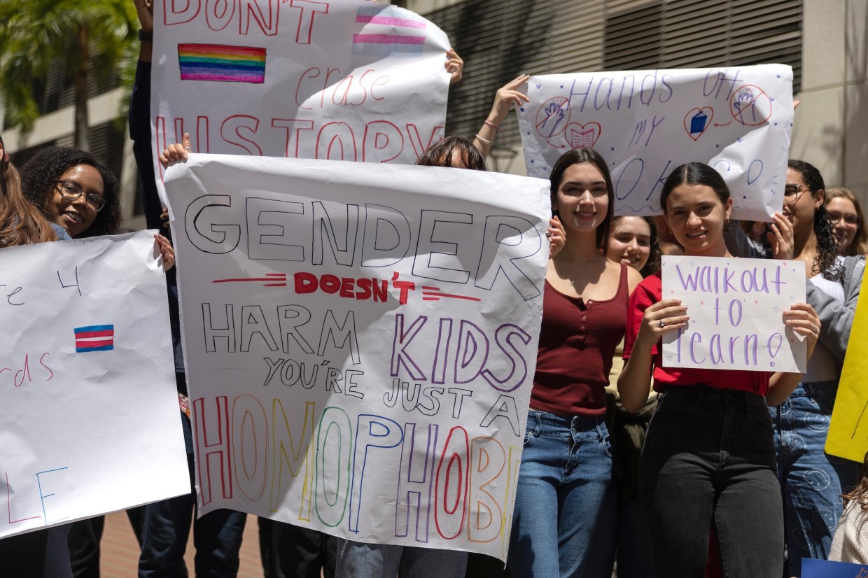 students hold up hand-drawn signs that read "Gender Doesn't Harm Kids You're Just a Homophobe," "Don't Erase History," "Walkout to Learn," and "Hands Off My Books"