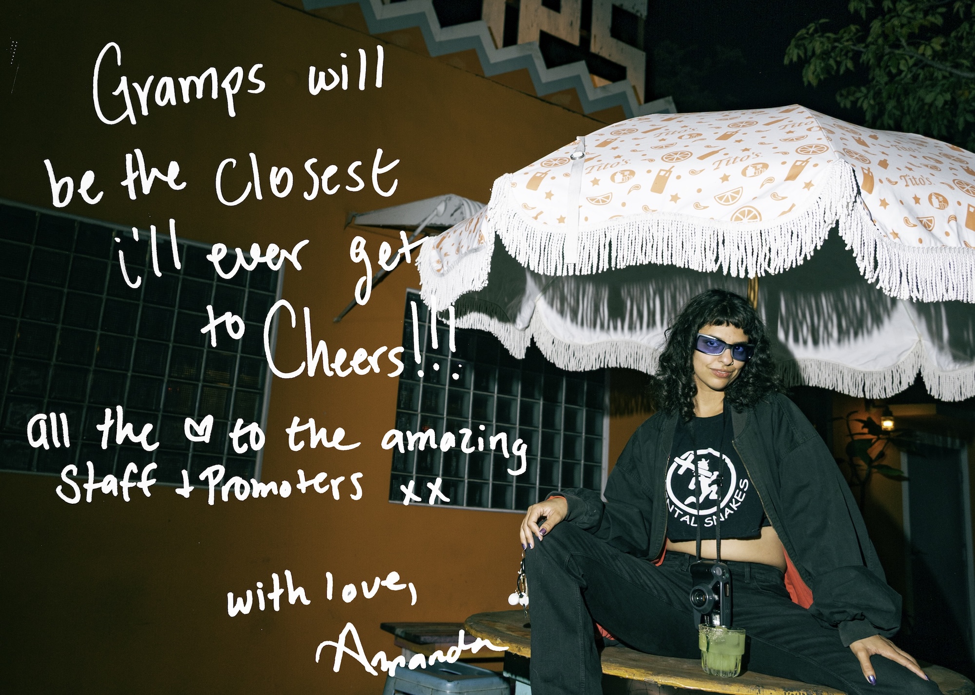 Photo of a woman sitting under an outdoor restaurant umbrella in front of an orange building. Text written on the photo reads, "Gramps will be the closest I'll ever get to Cheers! All the [heart symbol] to the amazing staff + promoters xx. With Love, Amanda"