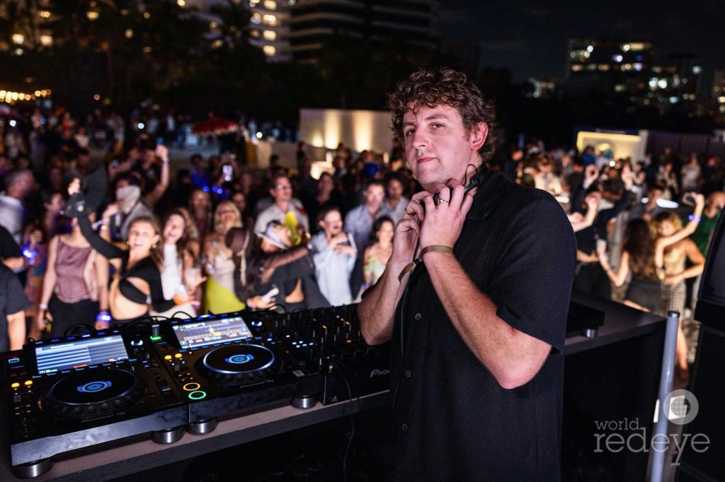 photo of a Jamie XX posing for a camera in front of a DJ booth with a crowd behind him
