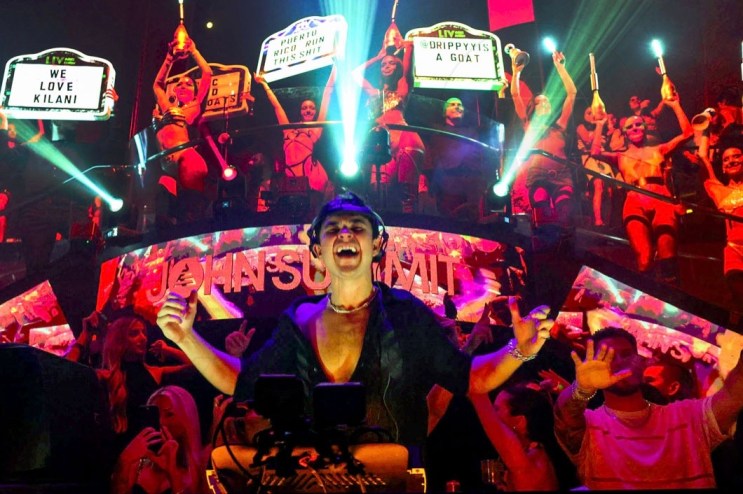 photo of DJ John Summit performing at a club with bottle girls holding up signs behind him