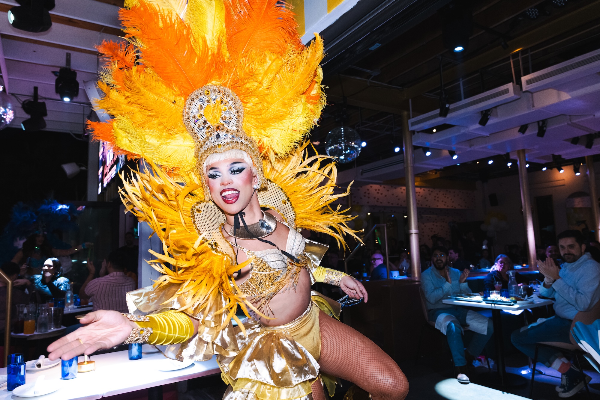 photo of a drag queen in a yellow showgirl outfit performing in a restaurant with diners in the background