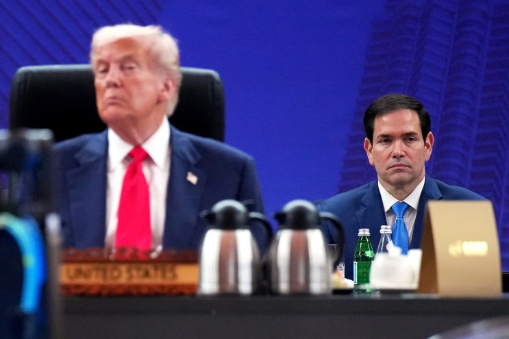 photo showing Donald Trump seated in a high-backed executive chair, out of focus in the foreground with a somewhat pompous expression, with Marco Rubio visible, in sharp focus, behind Trump's left shoulder, with a serious, somewhat sad expression. two insulated pitchers and two small san pellegrino bottles are visible in front of rubio