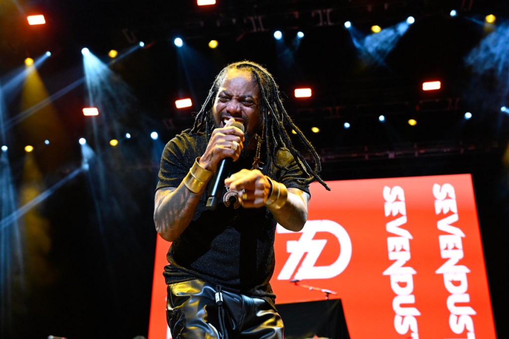 Photo of a man singing on stage wearing black clothes against an LED screen that reads Sevendust.
