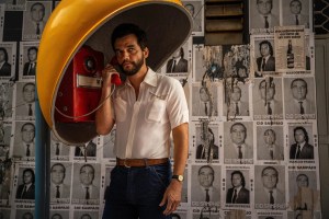 Photo of a man in a yellow, old-school telephone booth speaking on a red telephone. Behind him are black-and-white flyers with headshots plastered on a wall