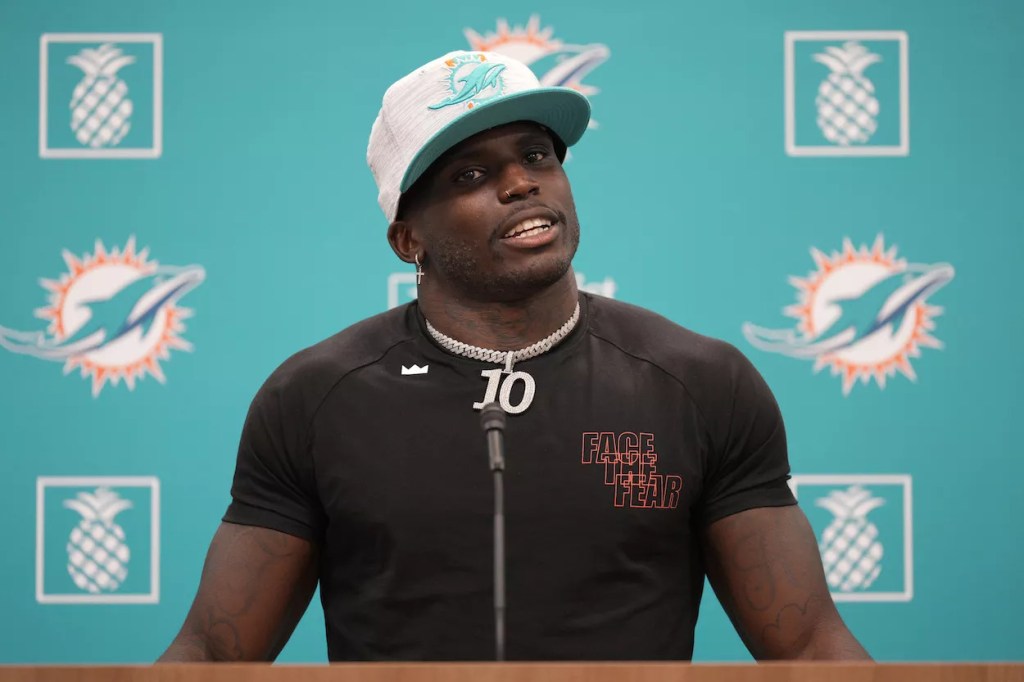 Tyreek Hill speaks at a press conference weating a white Dolphins hat and a fitted black t-shirt and a large silver chain.