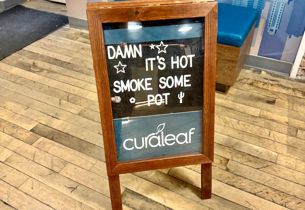 a sandwich board sign that reads "Damn It's Hot, Smoke Some Pot" with the Curaleaf logo underneath