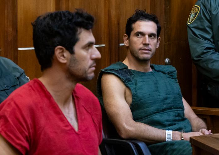 photo of a man in a red jumpsuit and another man wearing a navy kevlar vest inside a court room.