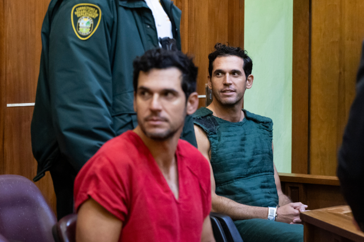 two handcuffed men, one in a red shirt and one in a navy kevlar vest, sit in a court room and look toward their right