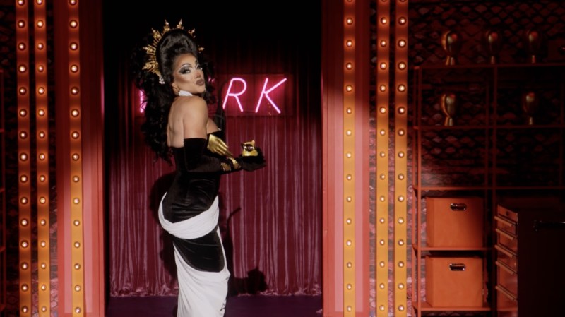 Photo of a drag queen wearing a black and white gown with a gold crown in front of a neon sign. Only the letters "R" and "K" are visible on the sign
