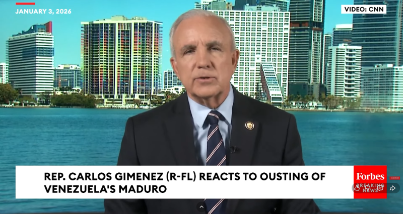 A man speaks on a TV broadcast with blue ocean and city skyline in the background.