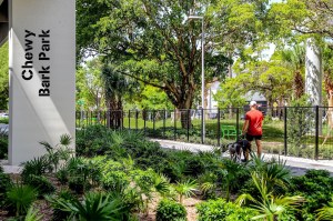 Photo of a person walking two dogs along a path under an overpass. A column below the overpass has a sign reading, "Chewy Bark Park"