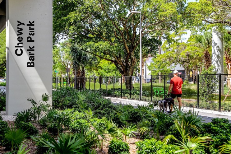 Photo of a person walking two dogs along a path under an overpass. A column below the overpass has a sign reading, "Chewy Bark Park"