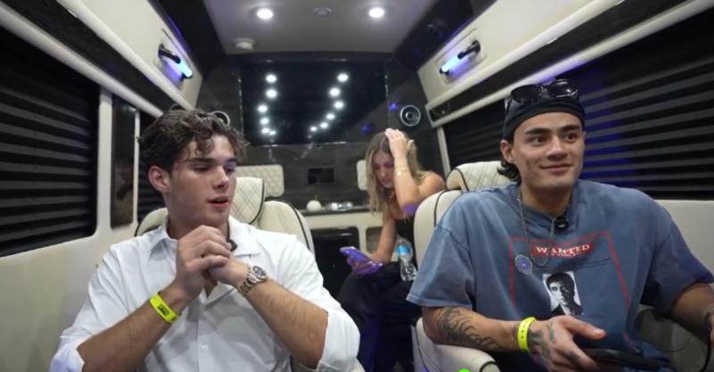 Clavicular sits on the left in a sprinter van. He is wearing a white-button down shirt. He is seated next to another man wearing a blue t-shirt. There is woman sitting behind them.