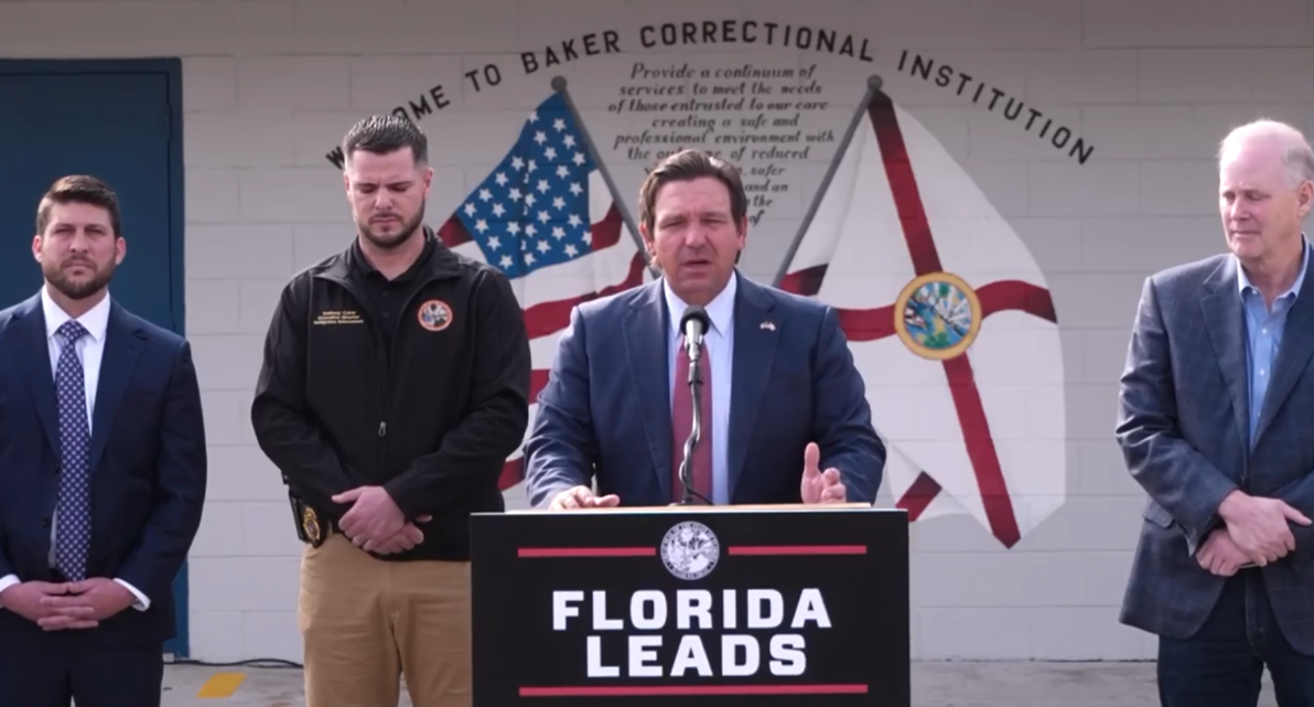 Ron DeSantis speaks at podium in front of the Baker Correctional Insitution. There are three men standing behind him. Two to his right and another on his left.